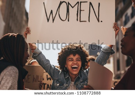 Laughing young woman holding a banner during a protest. Group of females activist protesting on road for women empowerment.