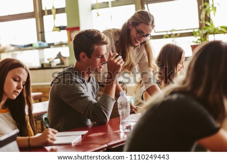 Female professor talking to one of students at lecture and looking at his notebook. Teacher during her class with students sitting at the desks.