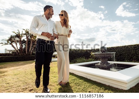 Stylish couple walking outdoors in lawn with a glass of wine. Man and woman looking at each other and walking together outdoors..