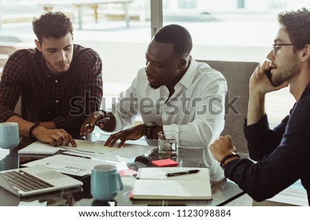 Businessmen discussing work sitting at conference table in office. Two men discussing work while another man is talking on mobile phone.