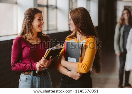 Two young women with book chatting while standing in college corridor. University students in corridor after the lecture.