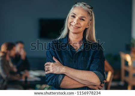 Portrait of confident senior business woman standing in office with her arms crossed. Mature female in office with colleagues discussing work at the back.