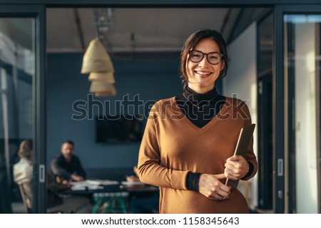 Portrait of positive female executive holding a laptop standing in office with colleagues working in background. Smiling business woman in casuals at office.