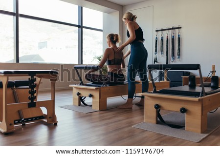 Fitness women doing pilates workout sitting on a pilates machine in a gym. Rear view of a trainer guiding a woman doing pilates workout at the gym.