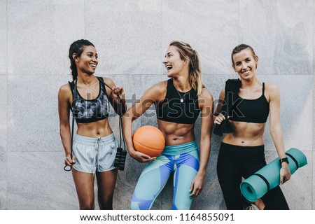 Happy women in fitness clothes relaxing and having fun standing against a wall. Fitness women carrying skipping rope basketball and workout mat standing after workout.