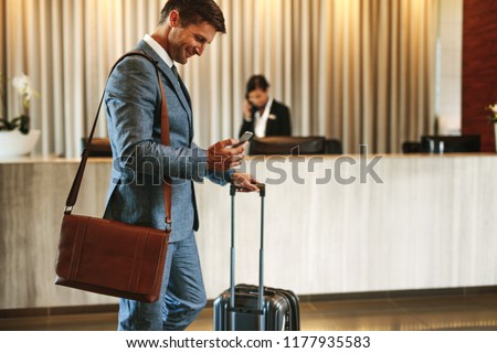 Businessman standing in hotel lobby with suitcase and using his mobile phone. Male business traveler in hotel hallway with smartphone and luggage.
