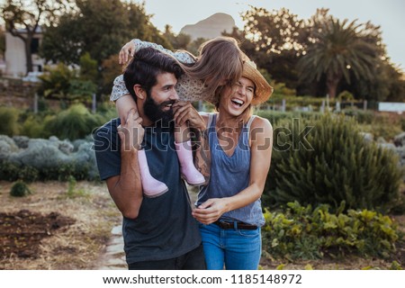 Beard man carrying little girl on shoulder with woman laughing at the farm. Beautiful family of three having fun at their farm.