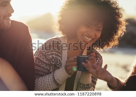 Young woman with coffee cup laughing while sitting with friends at the beach. Cheerful friends at the beach at sunset.