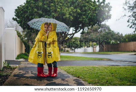 Two adorable little girls standing outdoors on a rainy day under an umbrella wearing raincoats. Twin sisters in waterproof coats standing together under umbrella outside.