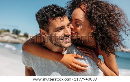 Smiling woman piggy riding on her boyfriend and kissing him. Man carrying his girlfriend on his back outdoors.