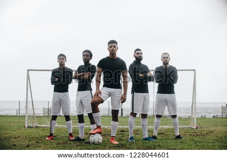 Five football players standing in a formation in front of goalpost. Soccer player standing with one foot on ball with teammates standing behind him with arms crossed.