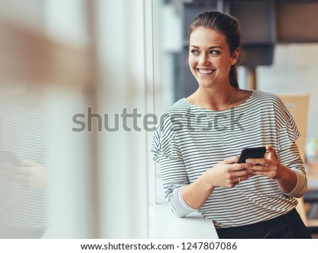 Cheerful woman entrepreneur standing beside a window and looking out. Businesswoman standing in office with a mobile phone in hand.