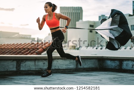 Female athlete doing drag running on rooftop. Side view of a fitness woman running on terrace of a building with a resistance parachute tied to her waist.