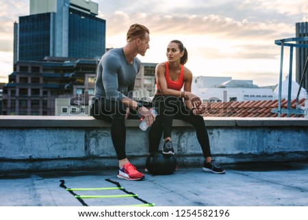 Man and woman in fitness clothes relaxing on the rooftop during workout. Fitness couple taking a break during workout sitting together on the fence of a terrace and talking.