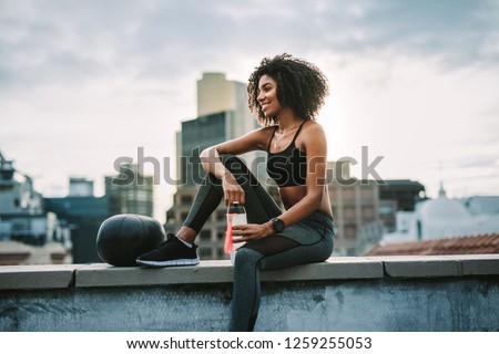 Side view of a woman athlete sitting on rooftop fence with a medicine ball drinking water after workout. Smiling woman in fitness clothes relaxing after workout sitting on rooftop.