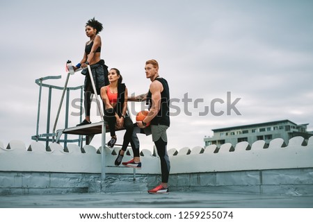 Man holding a basketball standing with two female athletes on rooftop stairs after fitness training. Low angle view of three fitness people relaxing after workout near rooftop stairs and looking away.