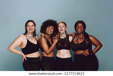Portrait of group of women posing together in sportswear against a gray background. Multiracial females with different size standing together looking at camera and smiling.