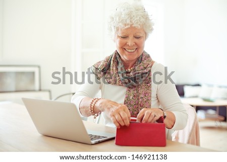 Cheerful old woman in front of laptop getting something from her wallet