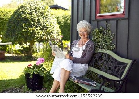 Relaxed elder woman sitting on a bench in backyard garden reading a newspaper looking at camera and smiling