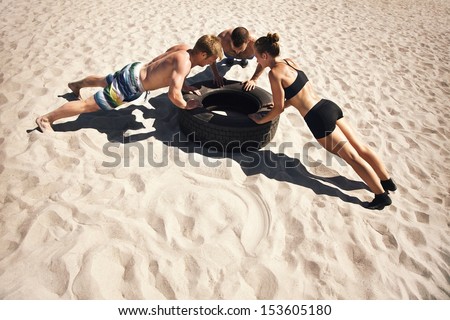 Small group of people doing push-ups on tire. Young athletes working out on beach during a hot summer day.