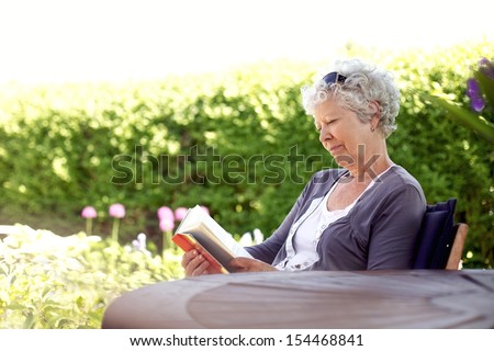 Senior woman reading a book sitting outside on a chair at the garden. Elder woman reading novel at her backward garden