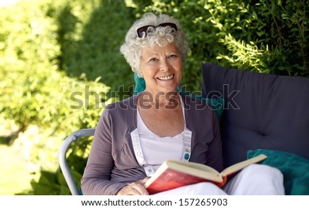 Elder Woman Sitting On A Chair In Backyard Garden Holding A Book And Looking At Camera Smiling
