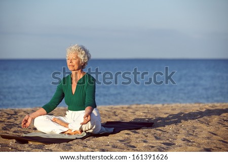 Image of old woman sitting on exercise mat practicing yoga on the beach. Senior caucasian woman meditating in lotus pose - Outdoors
