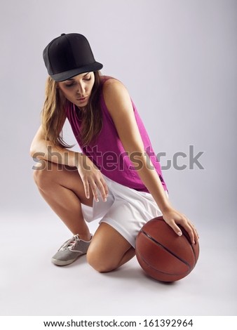 Image of attractive young woman wearing cap with a basketball sitting on floor. Female basketball player with ball on grey background