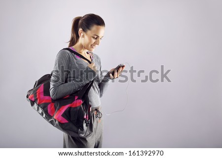 Good looking female athlete with a sports bag listening to music on her mobile phone Fitness woman in sports clothing going to gym on grey background Good looking female athlete with a sports bag listening to music on her mobile phone Fitness woman in sports clothing going to gym on grey background