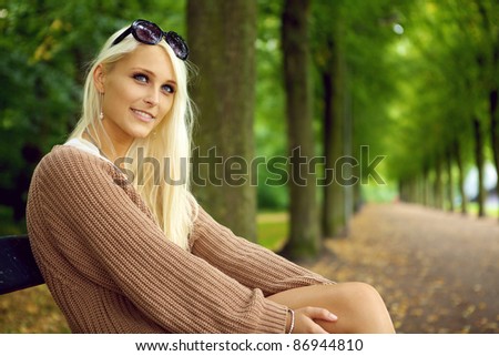 An attentive sexy young blonde woman in a tan jersey sits on a park bench looking upwards with empty tree lined avenue behind.