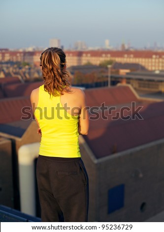 A young woman about to participate in parkour stands with her back to the camera on a high urban rooftop looking out over the city