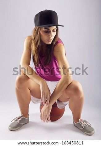 Portrait of attractive young female basketball player sitting on ball. Beautiful woman wearing cap with a basketball over grey background