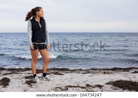 Full length image of beautiful young woman standing on beach looking away. Healthy female athlete relaxing after a run on sea shore. Caucasian young woman on beach looking at copyspace