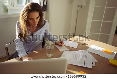 Pretty young businesswoman sitting at desk writing notes from a laptop at home. Beautiful caucasian woman working from home office.