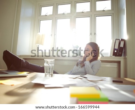 Young Woman Sitting At Her Table With Legs On Desk Talking On Mobile Phone. Caucasian Businesswoman Using Cell Phone While Relaxing At Her Desk. Female Working From Home Office.