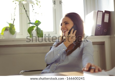 Pretty young woman talking on mobile phone while sitting at her desk. Beautiful businesswoman working from home office.
