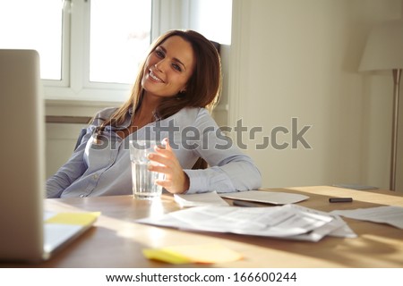 Pretty businesswoman sitting at her desk with a glass of water looking at camera smiling. Confident young caucasian woman working from home office.