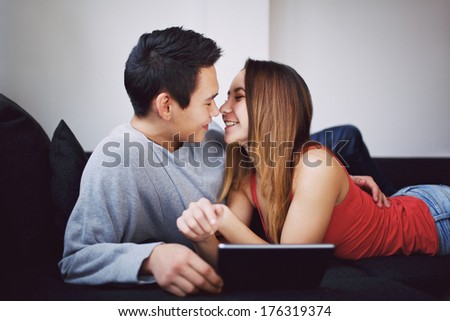 Romantic teenage couple on couch looking at each other with digital tablet. Young couple in lover about to kiss each other on sofa at home. Mixed race man and woman with tablet PC indoors.
