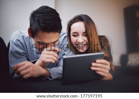 Happy Young Man And Woman Using Tablet Pc. Mixed Race Teenage Couple Using Digital Tablet Smiling While Lying On Sofa At Home.