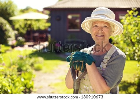 Portrait of senior woman wearing hat with gardening tools outdoors. Elder woman standing with shovel in her backyard garden