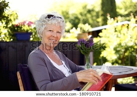 Senior woman sitting on a chair in backyard garden holding a book and looking at camera smiling