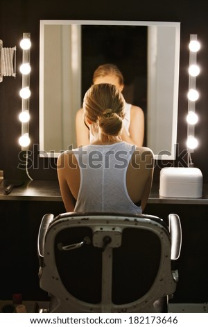 Rear view image of young woman sitting in front of mirror illuminated with light bulbs. Caucasian actress applying makeup backstage.