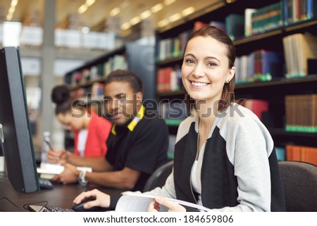 Beautiful young caucasian student sitting at table with computer looking at camera smiling. Young university students researching information on computer for their studies.