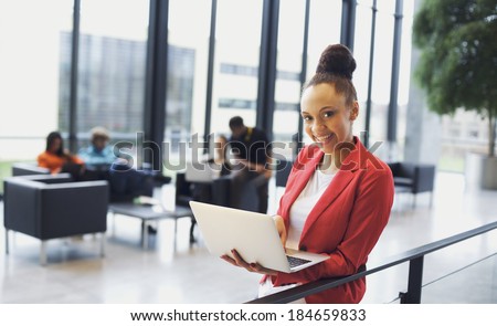 Beautiful young woman with a laptop in modern office. African american businesswoman standing by a railing with people working in background.