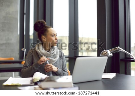 Young african american woman holding water bottle and looking away while studying in library. Female student sitting at table with laptop and books for study. Taking a break.