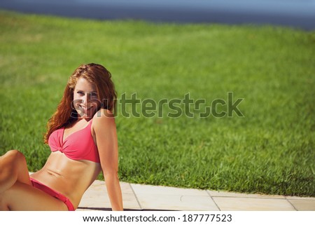 Pretty young woman in bikini sitting on floor along the grass lawn. Caucasian female model enjoying a sunbath on a hot summer day.