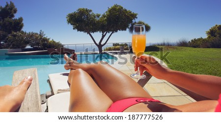 Young lady in bikini holding orange juice glass while sitting on a lounge chair along the poolside on a sunny day. Panorama view.