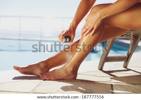 Woman sitting on deck chair by the swimming pool spraying suntan lotion onto her legs