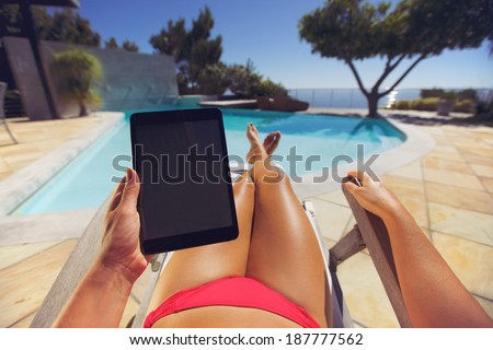 Young woman relaxing on a lounge chair using a tablet PC near the pool. User POV. Female model sitting on a deckchair holding digital tablet.