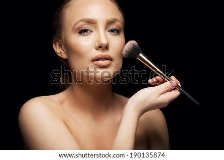 Beautiful young woman applying foundation on her face with a makeup brush isolated on black background. Attractive young caucasian model.